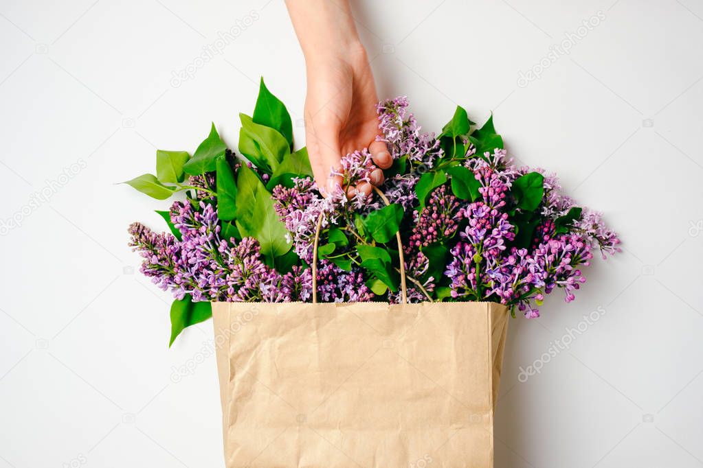 Women's hand holding craft paper package with lilac flowers over white background. Creative flat lay composition, top view, overhead. Spring sale, natural beauty, organic cosmetic shop market banner