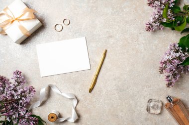 Düğün aksesuarları, boş kağıt kartı, altın kalem, parfüm şişesi, kurdele, yüzük ve bahar leylak çiçekleri ile kadınsı masa. Düğün konsepti, davetiye kartı mock up, düz yatıyordu, üst görünüm.