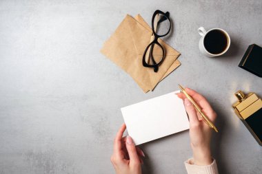 Düz boş kağıt kart, gözlük, kraft zarf, kahve fincanı, parfüm şişesi tutan kadın eli ile modern minimal ev çalışma alanı yatıyordu. Üst görünüm kadınsı masa, genel merkez, iş günlüğü kahraman.