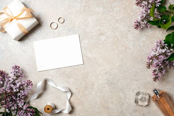 Feminine desk with wedding accessories, blank paper card, rings and spring lilac flowers. Wedding concept, invitation card mock up, flat lay, top view.