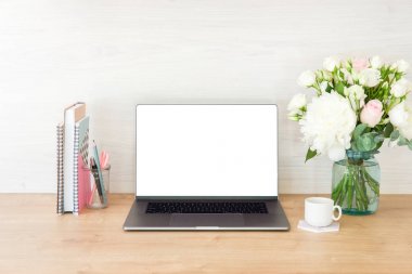 Boş beyaz ekranlı dizüstü bilgisayar, kadınsı ofis malzemeleri, çiçekler, kitaplar, kırtasiye malzemeleri, ahşap masa üzerinde kahve fincanı ile çalışma alanı.