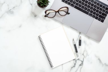Dizüstü bilgisayar, kağıt not defteri, gözlük, kırtasiye, beyaz mermer yüzey üzerinde sulu bitki ile Ofis masası masa. Düz yatıyordu, üst görünüm kadınsı arka plan. İş kadını çalışma alanı