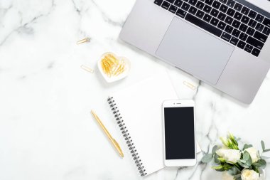 Düz yatıyordu, üst görünüm modern ofis masa masası. Dizüstü bilgisayar, boş ekranlı akıllı telefon, günlük, çiçekler ve mermer arka plan üzerinde altın kırtasiye ile çalışma alanı. İş kadını işyeri konsepti