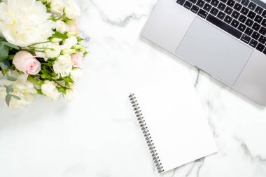 Düz yatıyordu ev ofis masası. Dizüstü bilgisayar, çiçekler, beyaz mermer yüzey üzerinde kağıt not defteri ile Kadın çalışma alanı. Üst görünüm kadınsı arka plan. İş kadını işyeri konsepti.