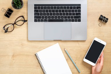 Dizüstü bilgisayar ile Ahşap ofis masası masa, siyah ekranlı el tutan akıllı telefon, sulu bitki, boş kağıt not defteri, kalem, gözlük, klips, kırtasiye. Kopyalama alanı ile üst görünüm, düz döşeme.