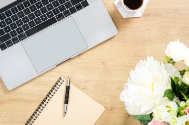 Dizüstü bilgisayar, çiçek buketi, kahve fincanı, kağıt not defteri, kalem ile ahşap ofis masası masa. Düz yatıyordu, üst görünüm şık kadın çalışma alanı. Kadınev ofis masası konsepti.