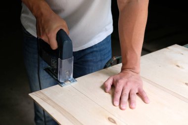 Marangoz marangozluk atölyesinde elektrikli jig testere makinesi ile bir kontrplak levha biçiyor. Yapbozla el ele. Marangozluk konsepti.