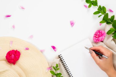 Düz yatıyordu, üst görünüm ofis masa masası. Hasır şapkalı çalışma alanı, boş kağıt not defteri, insan eli tutan kalem, pembe gül çiçeği tomurcukları ve beyaz arka plan üzerine yaprak.