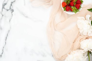 Üst görünüm pastel bej eşarp, mermer arka plan üzerinde çilek ve beyaz şakayık çiçekler kase. Trendy kadınsı aksesuarları ile minimal düz yatıyordu stil kompozisyon. Güzellik veya moda günlüğü afiş mockup