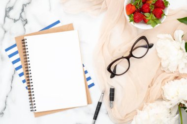 Üst görünüm boş kağıt not defteri, pastel bej eşarp, çilek kase, mürekkep kalem ve mermer arka plan üzerinde beyaz şakayık çiçekler. Trendy kadınsı aksesuarları ile Flatlay kompozisyon. Moda blog afiş