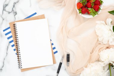 Boş kağıt not defteri, mürekkep kalem, çilek kase, pastel renkler eşarp ve mermer arka plan üzerinde beyaz şakayık çiçekler. Kadınsı aksesuarları ile minimal düz yatıyordu stil kompozisyon. Moda veya güzellik günlüğü