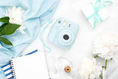 Üst te anlık fotoğraf kamerası, kağıt not defteri, beyaz şakayık çiçekler, mermer arka plan üzerinde mavi eşarp. Kadınsı aksesuarları ile minimal düz yatıyordu stil kompozisyon. Moda veya güzellik günlüğü afiş mockup.