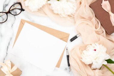 Üst görünüm boş düğün davetiyekartı ve beyaz şakayık çiçekler ve pastel bej eşarp ile mermer arka plan üzerinde zanaat kağıt zarf. Minimal flatlay tarzı kompozisyon, düğün ve evlilik kavramı