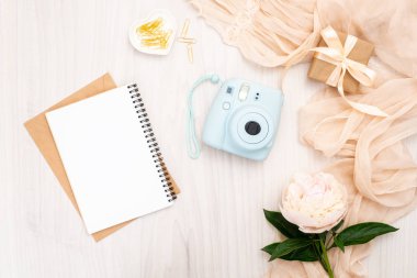 Rahat kadınsı ev ofis masası. Modern anlık kamera, bej eşarp, şakayık çiçekler, kağıt not defteri. Üst görünüm, ihale minimal düz döşeme tarzı kompozisyon. Kadın masası, moda blogger, güzellik teknolojisi