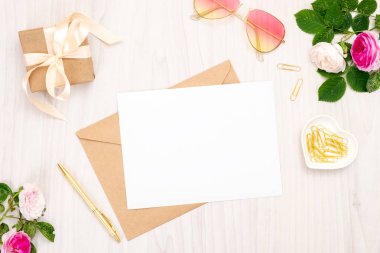 Düz yatıyordu kadın öğeleri: boş beyaz kağıt kartı ile zanaat zarf, gül çiçekleri, güneş gözlüğü, hediye kutusu, ahşap arka plan üzerinde altın klipler. Üst görünüm kadınsı aksesuarlar, güzellik günlüğü afiş mockup.