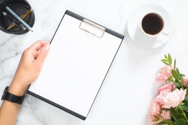 Kadın elinde boş kağıt pano ile Modern ev ofis masası çalışma alanı, pembe gül çiçek buketi, kahve fincanı, beyaz mermer taş arka plan üzerinde kırtasiye. Flatlay, üst görünüm.