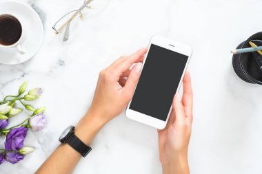 Kırtasiye, kahve fincanı, çiçek buketi, gözlük ile modern ev ofis masası çalışma alanı üzerinde siyah ekran mockup ile akıllı telefon tutan kadın eller. Düz döşeme, üst görünüm