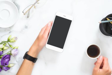 Kırtasiye, çiçek buketi, gözlük ile modern ev ofis masası çalışma alanı üzerinde siyah ekran mockup ve kahve fincan ile akıllı telefon tutan kadın eller. Düz döşeme, üst görünüm.