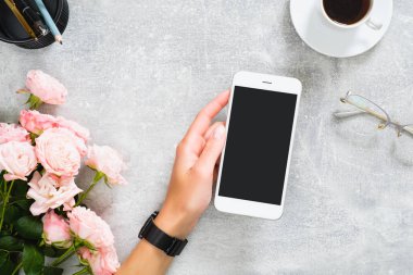 Boş ekran mockup ile akıllı telefon tutan kadın el. Gül çiçekleri, kahve fincanı, kırtasiye, beton taş arka plan üzerinde gözlük ile Kompozisyon. Kadınsı masa, güzellik teknolojisi konsepti.