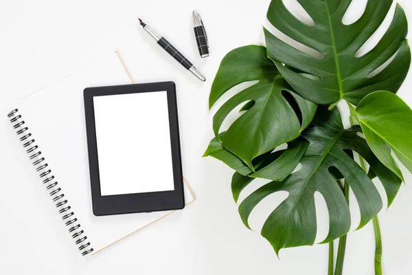Flat lay tropical jungle Monstera leaves, paper notebook, e-book reader on white background. Top view feminine diary, stationery and electronic device on home office desk.