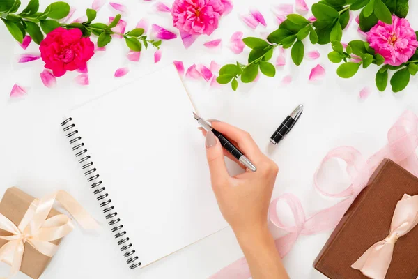 Женская рука пишет текст в бумажном блокноте дневник лежал на белом фоне с розовыми цветочными бутонами розы и подарочные коробки. Женское рабочее пространство, плоский уголок, вид сверху стильная концепция искусства . — стоковое фото