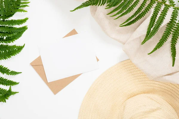 Summer vacation concept with blank paper card, craft paper envelope, feminine straw hat, tropical fern leaf on white background. Minimal flat lay style composition, top view, overhead.