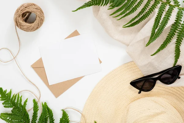 Summertime concept with craft paper envelope, blank postcard, straw hat, twine, tropical fern leves and sunglasses on white background. Flat lay, top view