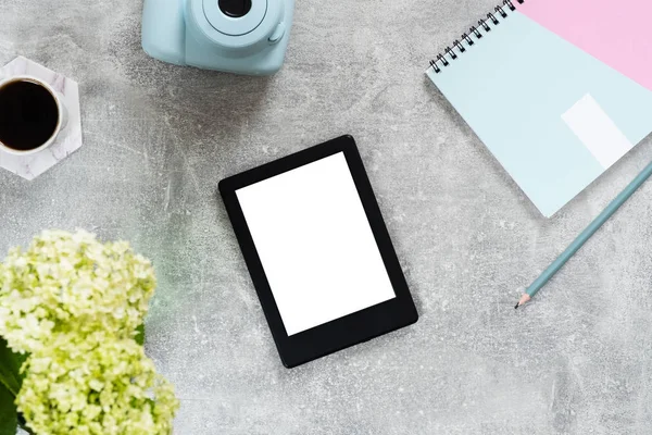 Kadınsı aksesuarları ve cihazları ile üst görünüm ev ofis masası. Düz boş beyaz ekran, kağıt not defteri, anlık film kamera, kahve fincanı ve beton yüzeyüzerinde çiçekler ile e kitap okuyucu yatıyordu.