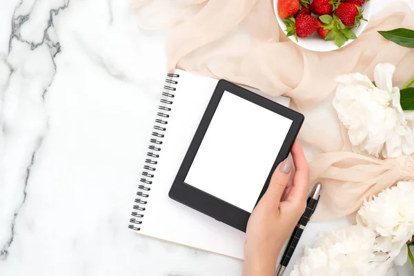 Top vista mão mulher segurando leitor de e-book em fundo de mármore com morango, cachecol de cor pastel e flores de peônia branca. Composição de estilo de leigos plana mínima. Moda ou beleza blog banner mockup . — Fotografia de Stock