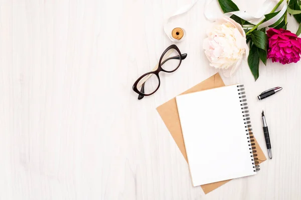 Flat lay home office desk. Female workspace with paper notebook, bouquet of pion flowers, golden accessories, glasses on wooden background. Top view feminine background. Fashion blog banner template.