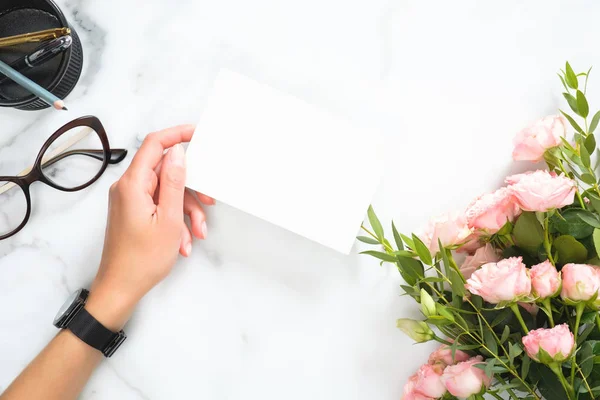 文房具付きのモダンなホームオフィスデスクワークスペースの上に白紙カードモックアップを持つ女性の手、バラの花の花束、眼鏡、フラットレイ、トップビュー. — ストック写真