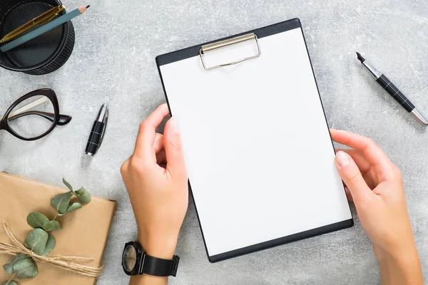 Kırtasiye, hediye kutusu, taş arka plan üzerinde gözlük ile şık kadınsı çalışma alanı üzerinde kadın ellerde Boş kağıt pano mockup. Düz yatıyordu, üstten bak. İş kavramı.