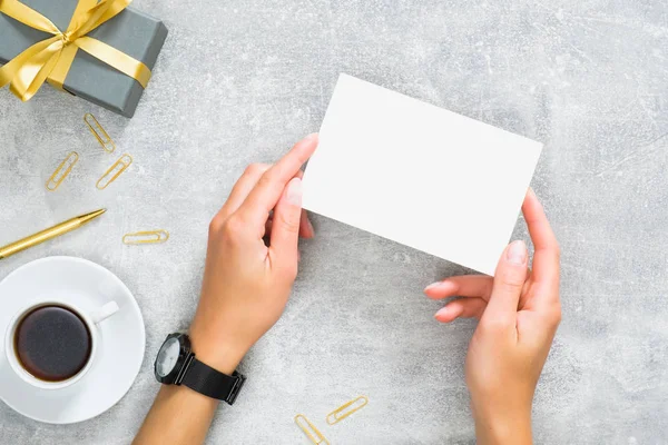 Hediye kutusu, altın kırtasiye ve kahve fincanı ile beton taş masada boş kağıt kart mockup tutan eller. Düz döşeme, üst görünüm, kopyalama alanı.