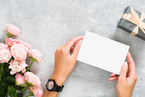Eller pembe gül çiçekleri ve hediye kutusu ile beton taş masada boş kağıt kart mockup tutan. Düz döşeme, üst görünüm, kopyalama alanı.