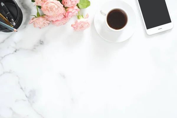 Flat lay, top view office table desk. Feminine desk workspace frame with coffee cup, floral bouquet, smartphone and stationery on white marble background.