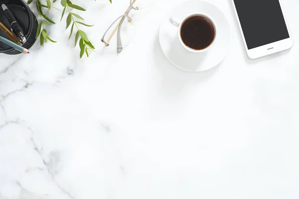 Flat Lay, topputsikt kontorsbord. Feminint skrivbord arbetsyta ram med kaffekopp, Glasögon, grön blommig filial, smartphone och brevpapper på vit marmor bakgrund. — Stockfoto