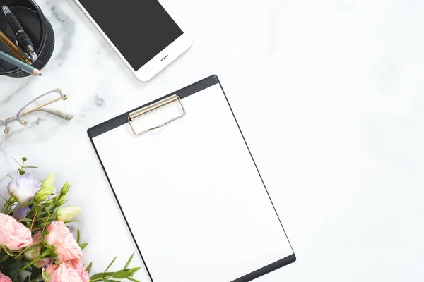 Espacio de trabajo moderno escritorio de oficina en casa con papel en blanco portapapeles maqueta, rosas flores, gafas, teléfono inteligente, papelería sobre fondo de mármol blanco. Pisos, vista superior . — Foto de Stock