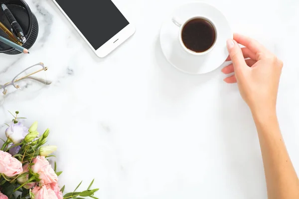 Kvinnliga handen håller kaffekopp. Komposition med rosor blommor, smartphone, Glasögon, brevpapper på marmor bakgrund. Flat Lay, uppifrån. Skönhets teknik, affärsidé. — Stockfoto