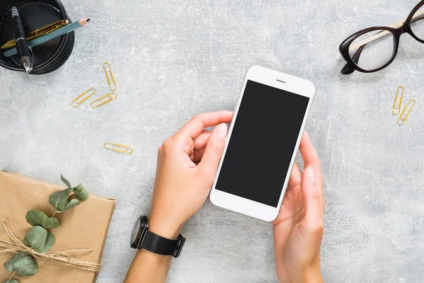 Manos de mujer sosteniendo smartphone con pantalla en blanco maqueta sobre elegante espacio de trabajo femenino con caja de regalo, papelería de color dorado, gafas. Plantilla de banner para aplicación móvil. Concepto de belleza y tecnología . — Foto de Stock