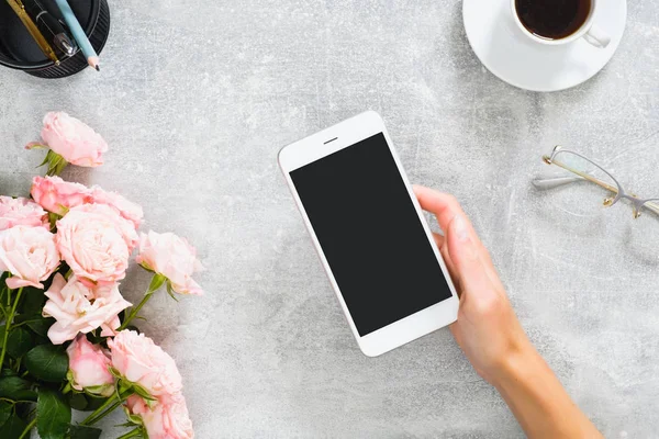 Mano femenina sosteniendo smartphone con pantalla en blanco maqueta. Composición con flores de rosas, taza de café, papelería, vasos sobre fondo de piedra de hormigón. Escritorio femenino, concepto de tecnología de belleza . — Foto de Stock