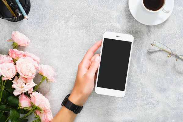 Female hand holding smartphone with blank screen mockup. Composition with roses flowers, coffee cup, stationery, glasses on concrete stone background. Feminine desk, beauty technology concept.