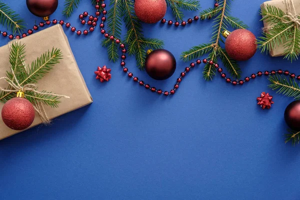 赤いクリスマスの装飾、モミの木の枝、クリスマスボール、青い背景にクラフト紙を包んだギフトボックス。クリスマス、冬、新年のコンセプト。フラットレイ、トップビュー、コピースペース — ストック写真