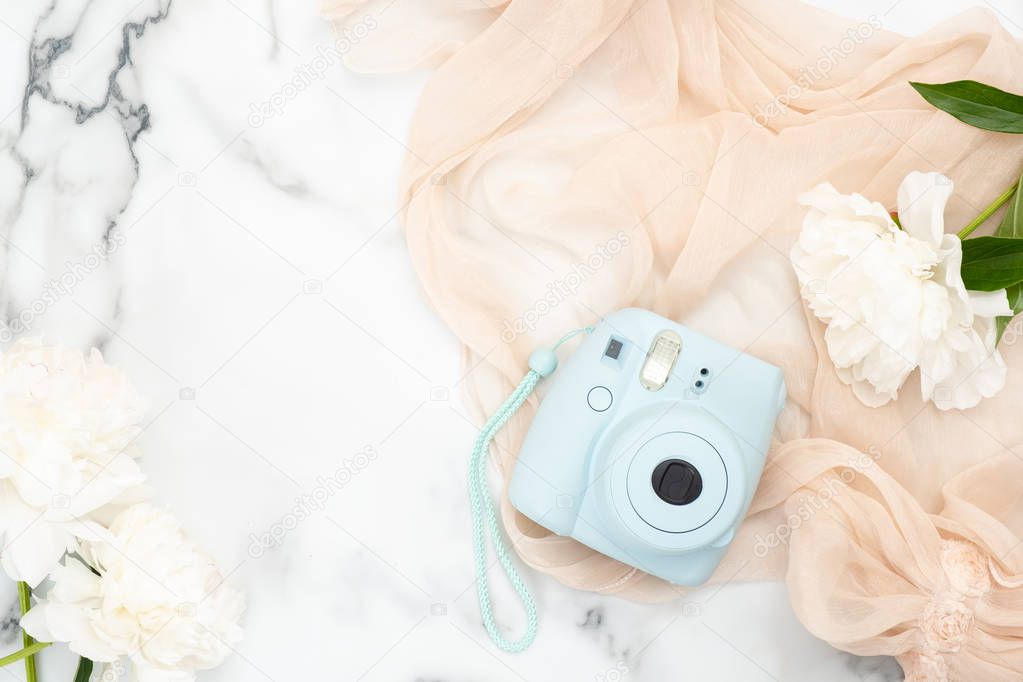 Top view instant film camera on marble background with white peony flowers and pastel color scarf. Minimal flat lay style composition with feminine accessories. Fashion or beauty blog banner mockup.