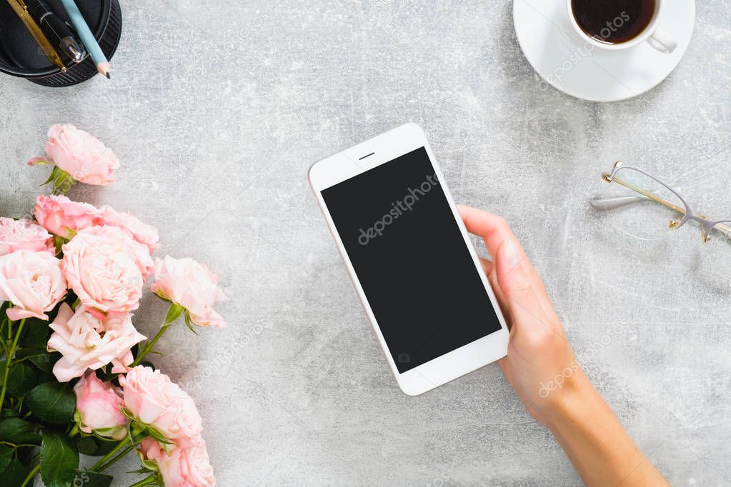 Female hand holding smartphone with blank screen mockup. Composition with roses flowers, coffee cup, stationery, glasses on concrete stone background. Feminine desk, beauty technology concept.