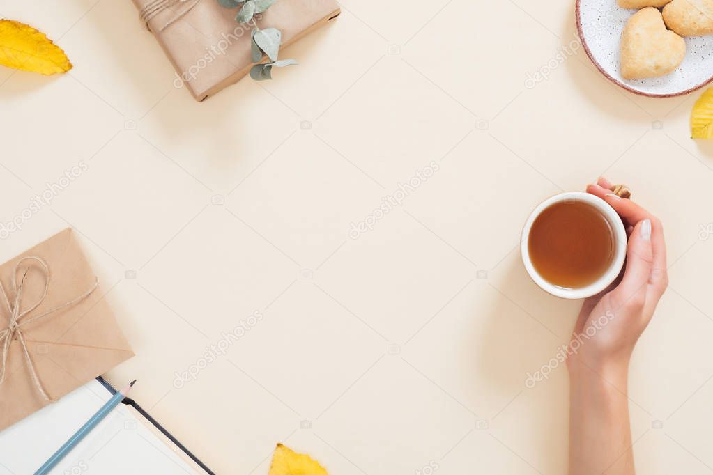 Woman hand holding cup of tea over autumn background with fallen leaves, book, cookies, gift box. Autumn composition, flat lay, top view, overhead Cozy home desktop, hygge concept.