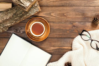 Çay fincanı ile Hygge tarzı flatlay kompozisyon, boş sayfaları ile kitap, beyaz örme eşarp, kadın gözlük, ahşap masa üzerinde yakacak odun. Sonbahar veya kış tatilleri kavramı. Düz döşeme, üst görünüm.