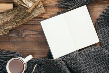 Boş kağıt defter, çay fincan, moda örme eşarp, ahşap arka plan üzerinde yakacak odun ile Hygge tarzı flatlay kompozisyon. Düz yatıyordu, üstten bak, tepede. Rahat sonbahar ev masası masası