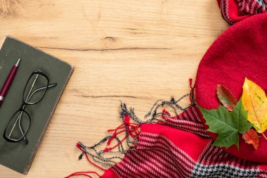 Hygge sonbahar konsepti. Örme atkı, kitap, gözlük, kırmızı kadınbesi, tahta masanın üzerine düşmüş yapraklar. Düz döşeme, üst görünüm, kopyalama alanı.