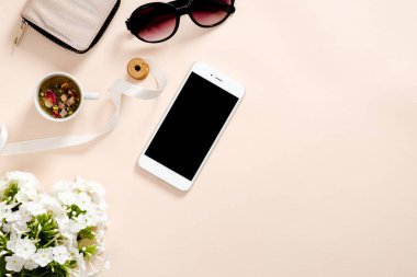 Kadınsı aksesuarları ile Flatlay kompozisyon, çay bardağı, akıllı telefon boş ekran mockup, güneş gözlüğü, pastel pembe arka plan papatya çiçekler. Kadınsı ev ofis masası konsepti. Düz döşeme, üst görünüm