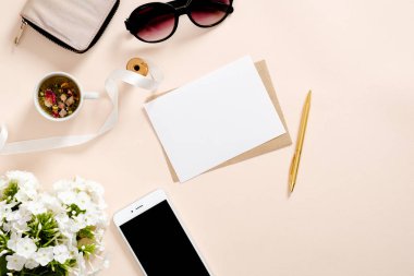 Kadınsı aksesuarları ile Flatlay kompozisyon, boş kağıt kart mockup, çay bardağı, akıllı telefon, güneş gözlüğü, pastel pembe arka plan papatya çiçekler. Kadınsı ev ofis masası konsepti. Düz döşeme, üst görünüm